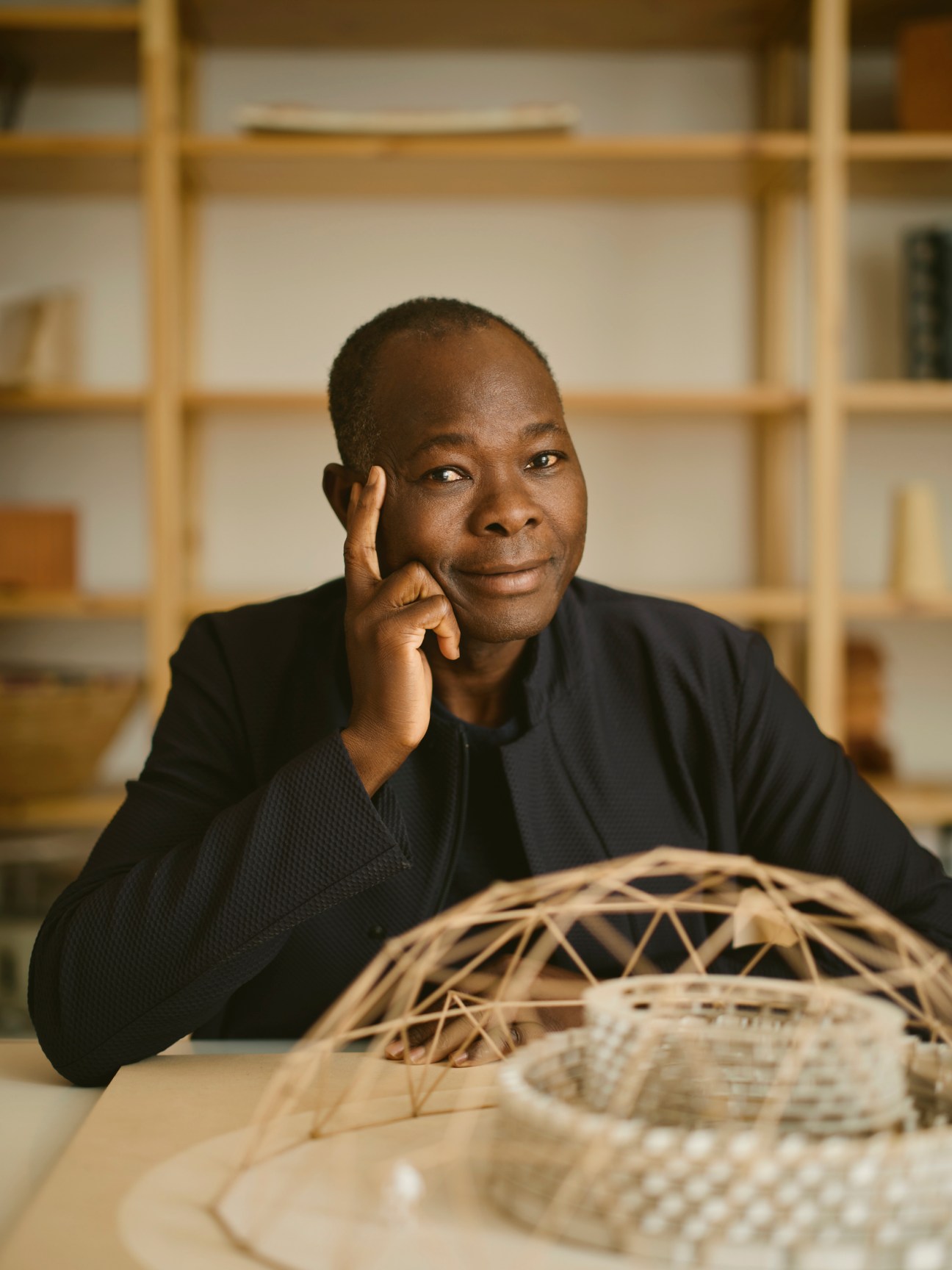 Diébédo Francis Kéré seated at his desk in his Berlin studio with a woven basket structure