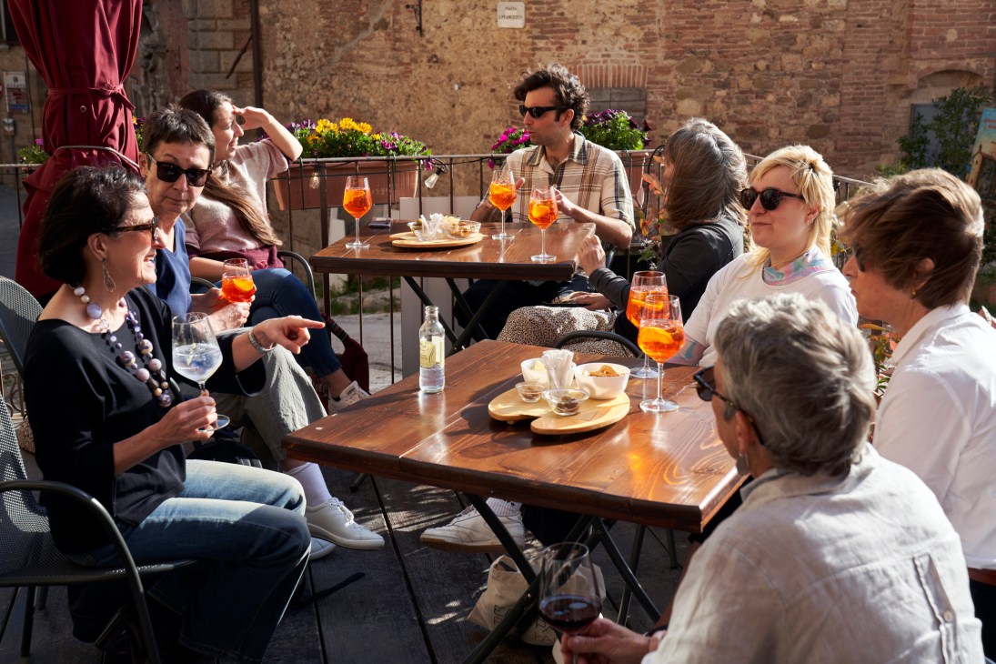 Group of people socializing at outdoor cafe tables with orange drinks and snacks in sunny courtyard