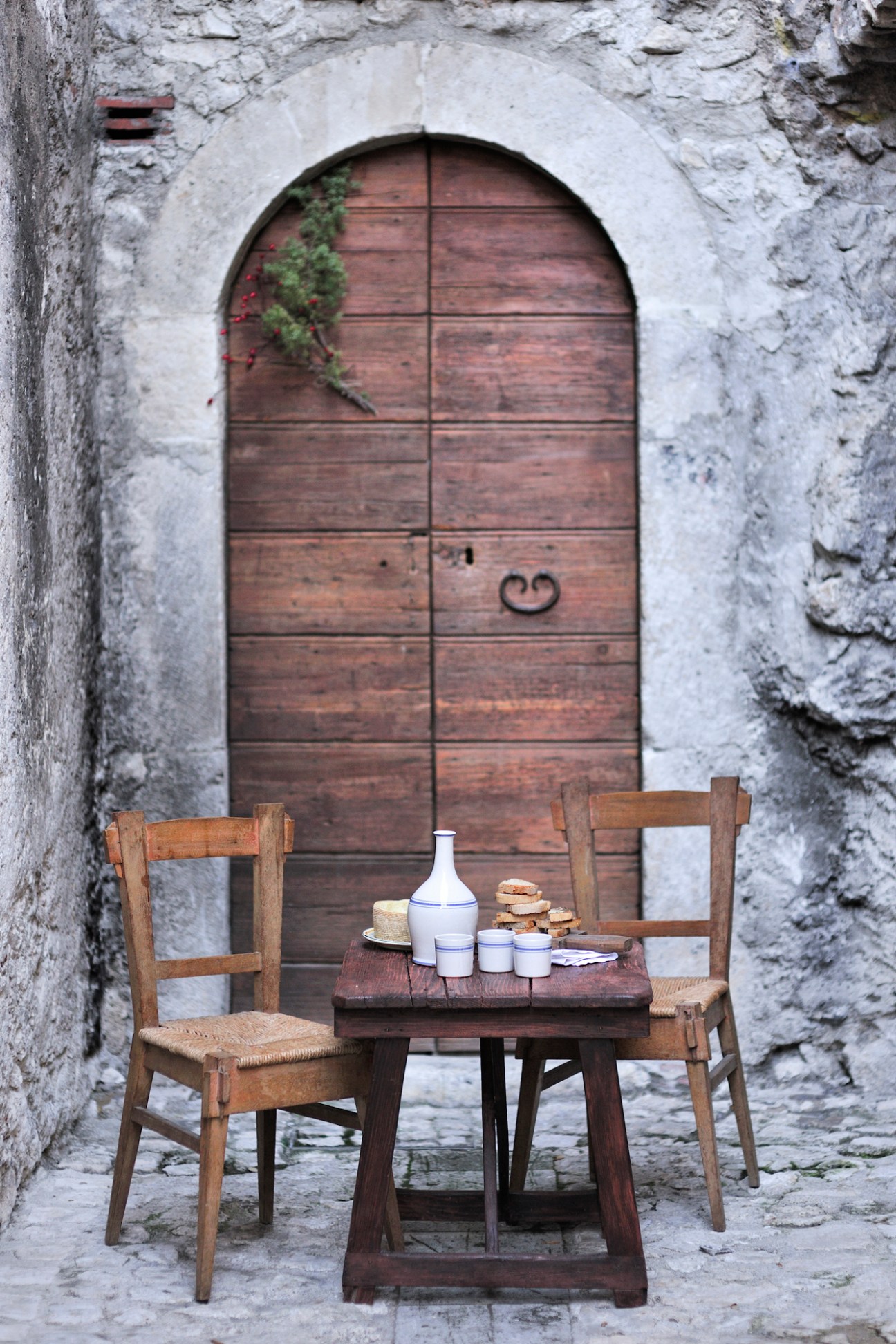 Rustic wooden table and chairs set with wine bottle and bread outside a weathered stone building with arched wooden door.