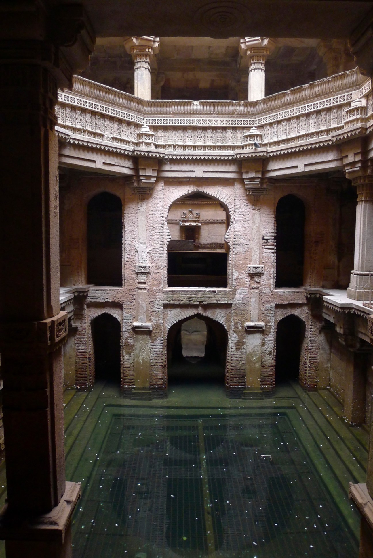 Multi-level stone stepwell with ornate carved pillars and arched openings containing green water at the bottom