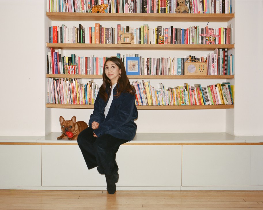 Alicia Kirby sits on white storage unit with built-in bookshelves behind, small dog beside her with red toy