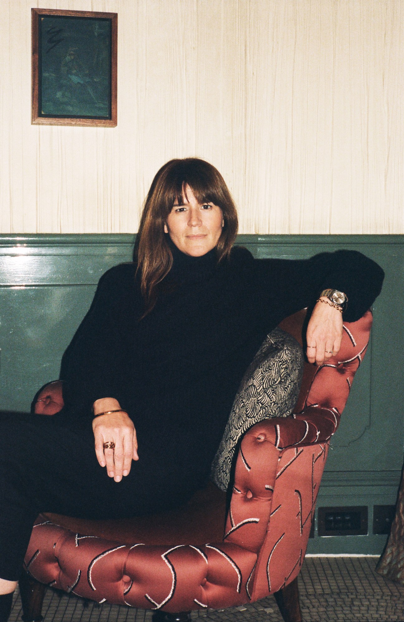 Rose Percy sitting on a green leather banquette wearing a black turtleneck and patterned pants