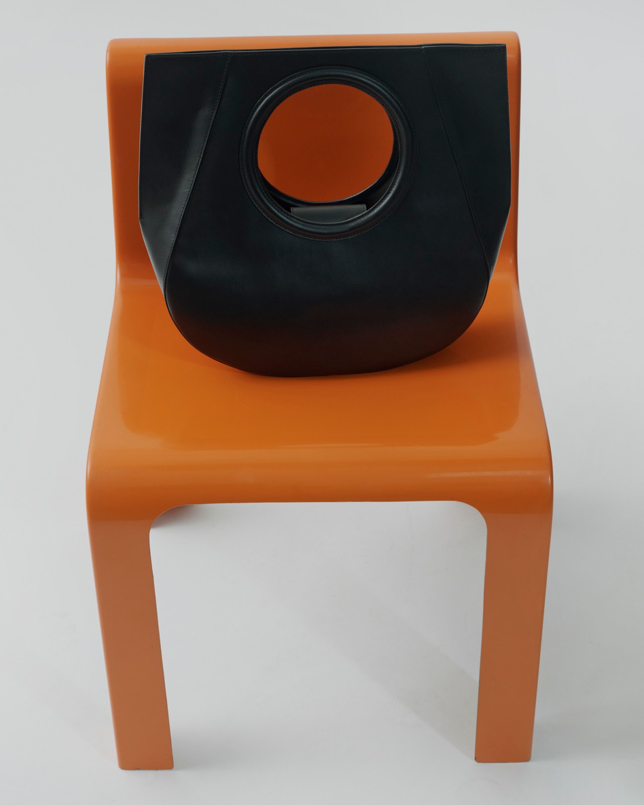 Black leather handbag by Hereu placed on orange plastic chair against white background
