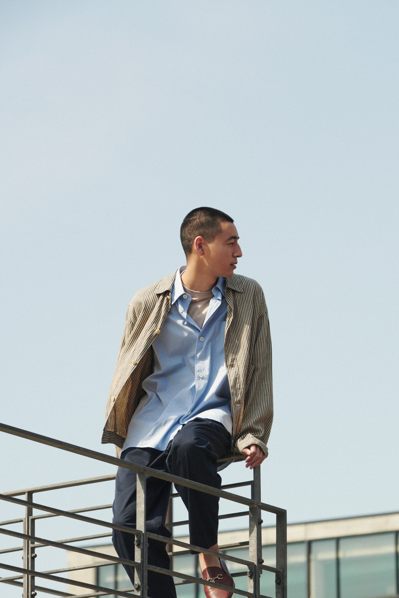 Man in striped cardigan, blue shirt and dark trousers sitting on metal railings against clear sky
