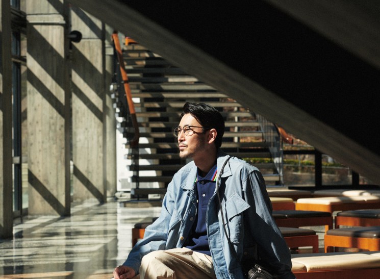 Asian man in glasses sitting on orange bench outdoors wearing layered clothing and holding a bag