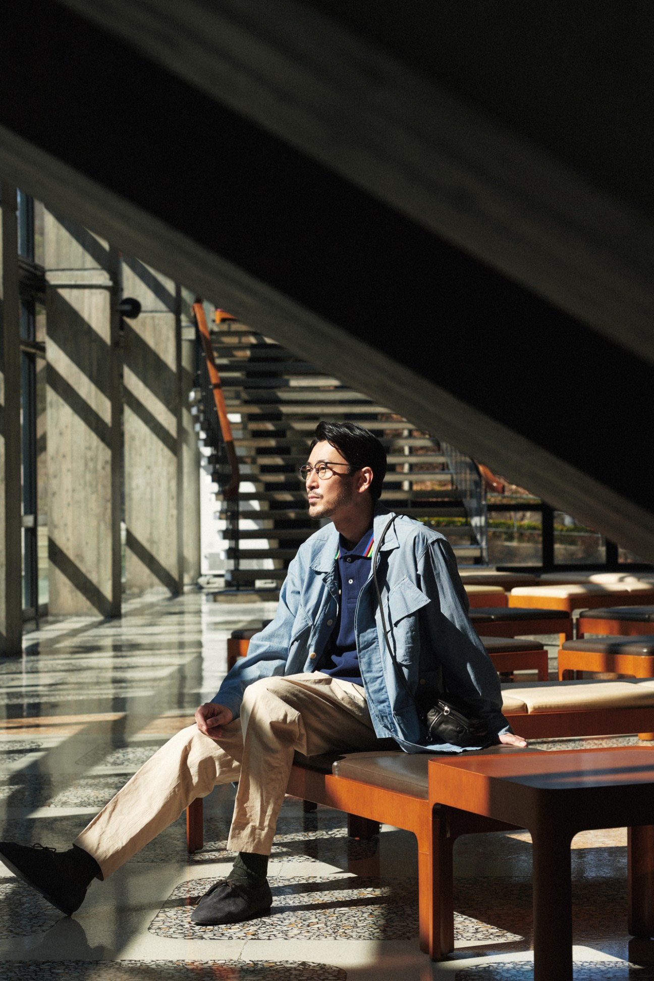 Asian man in glasses sitting on orange bench outdoors wearing layered clothing and holding a bag