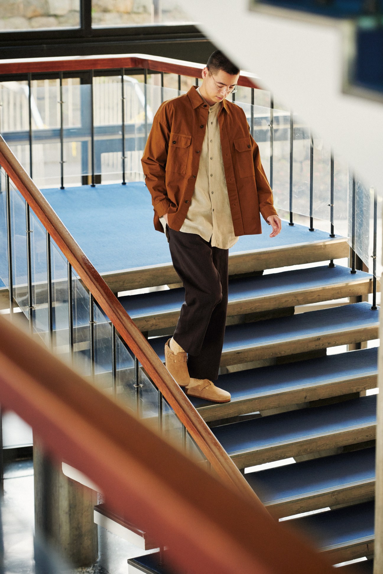 Man in brown overshirt and beige shirt walking down modern staircase with glass railings