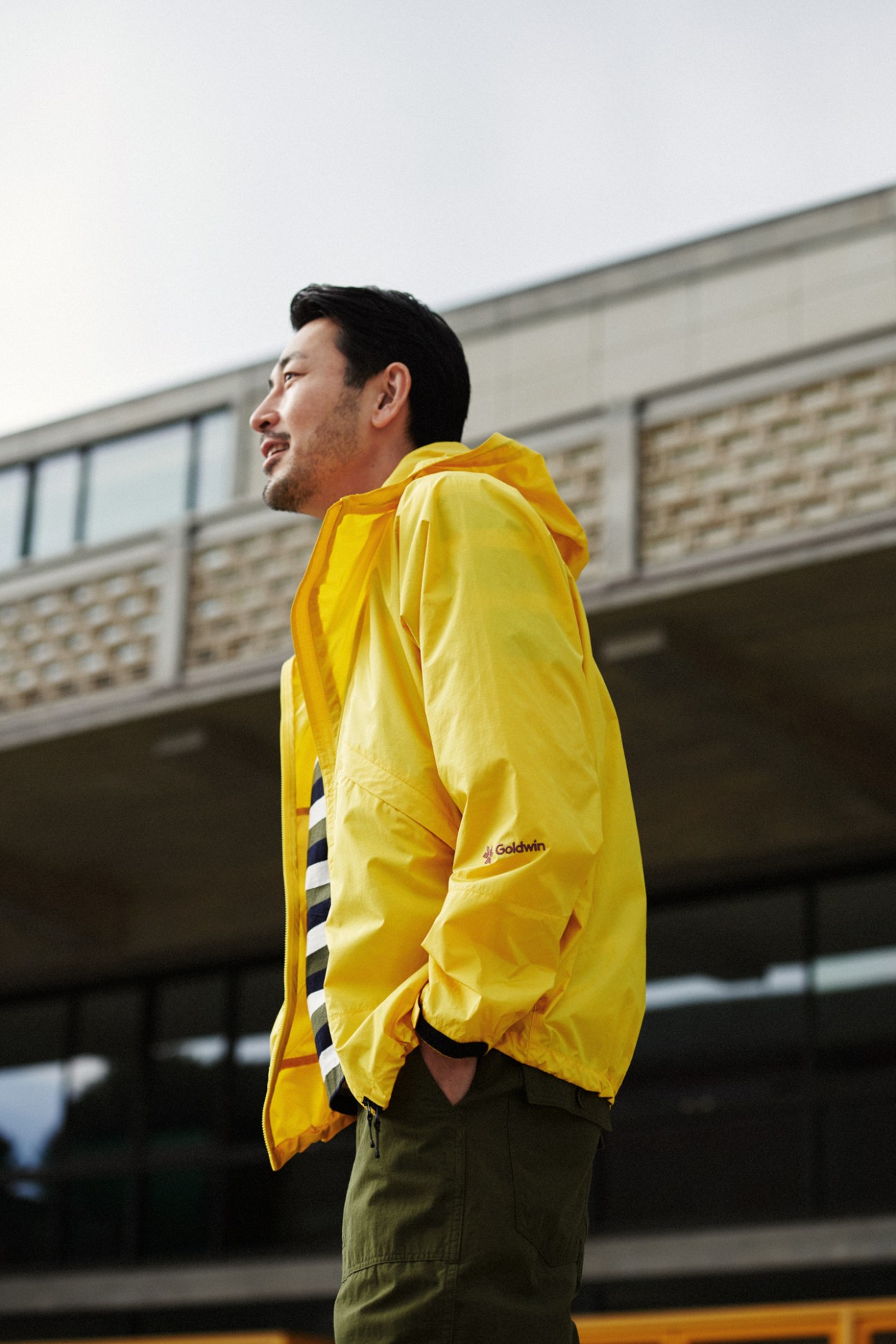 Man in yellow Goldwin jacket and olive trousers looking away in profile against urban building background