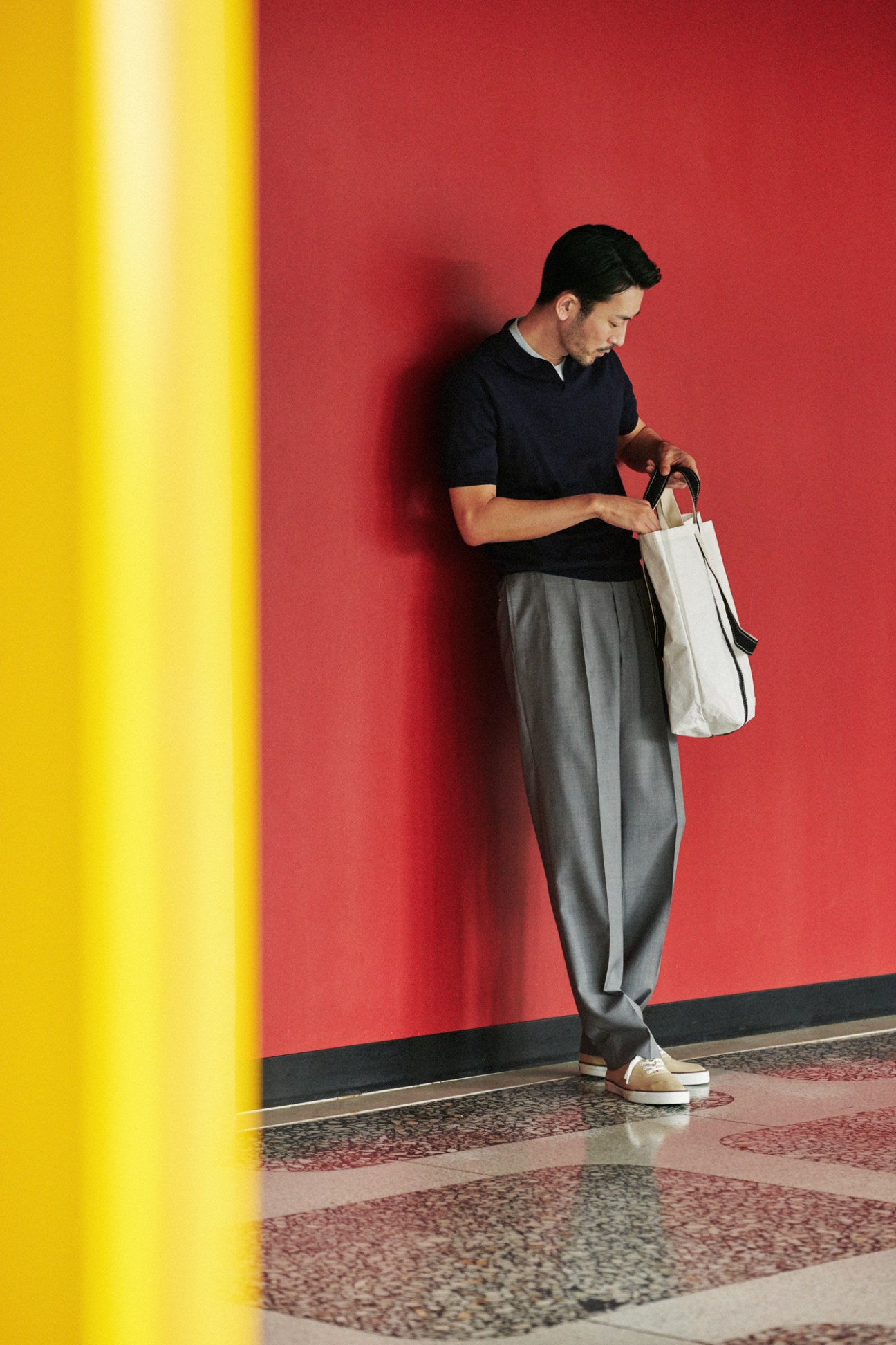 Man in dark polo shirt and grey trousers holding white bag against red wall
