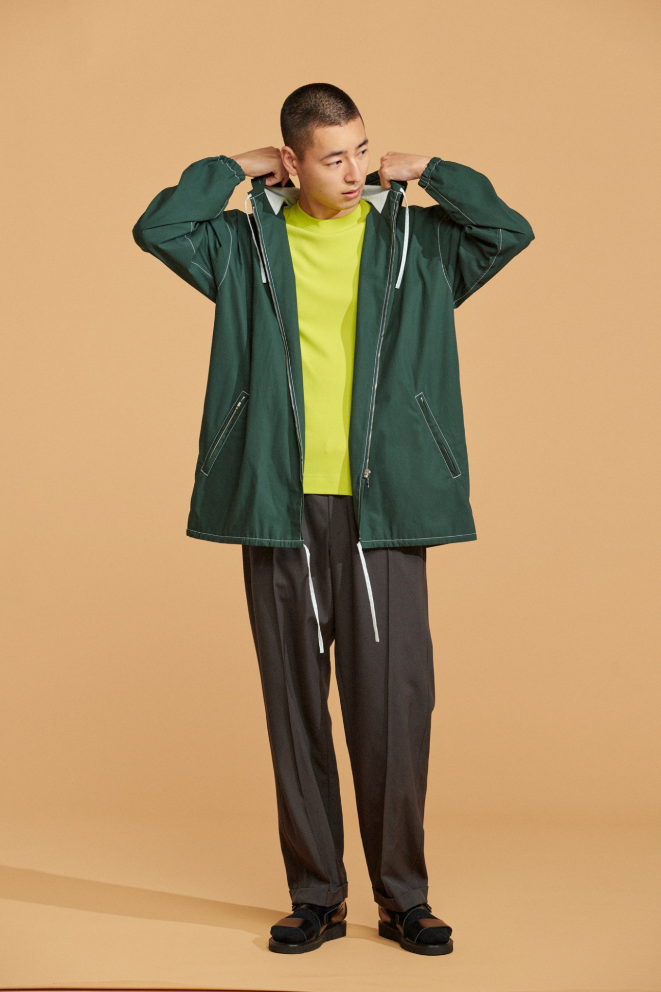 Model wearing green zip-up hoodie jacket over yellow t-shirt with dark trousers and black sandals