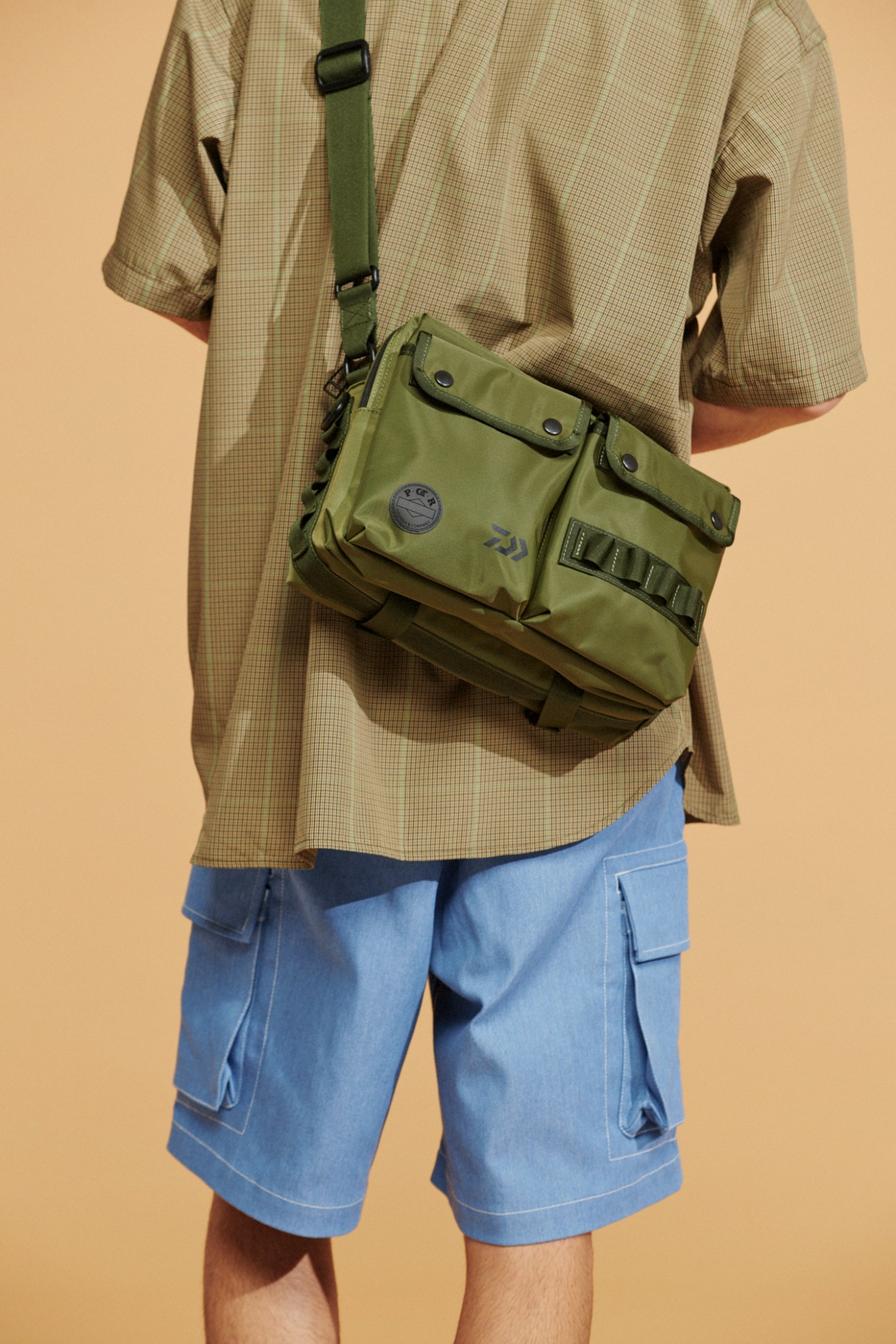 Model wearing olive green crossbody bag, checkered shirt and blue shorts against beige background