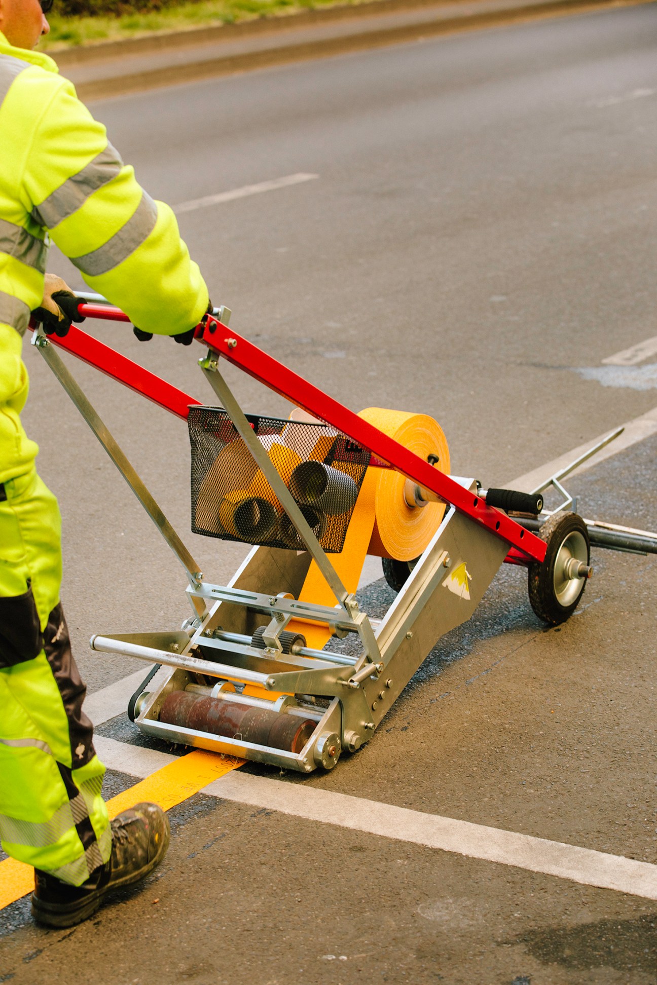 Worker in high-visibility jacket operating road marking equipment on asphalt street.