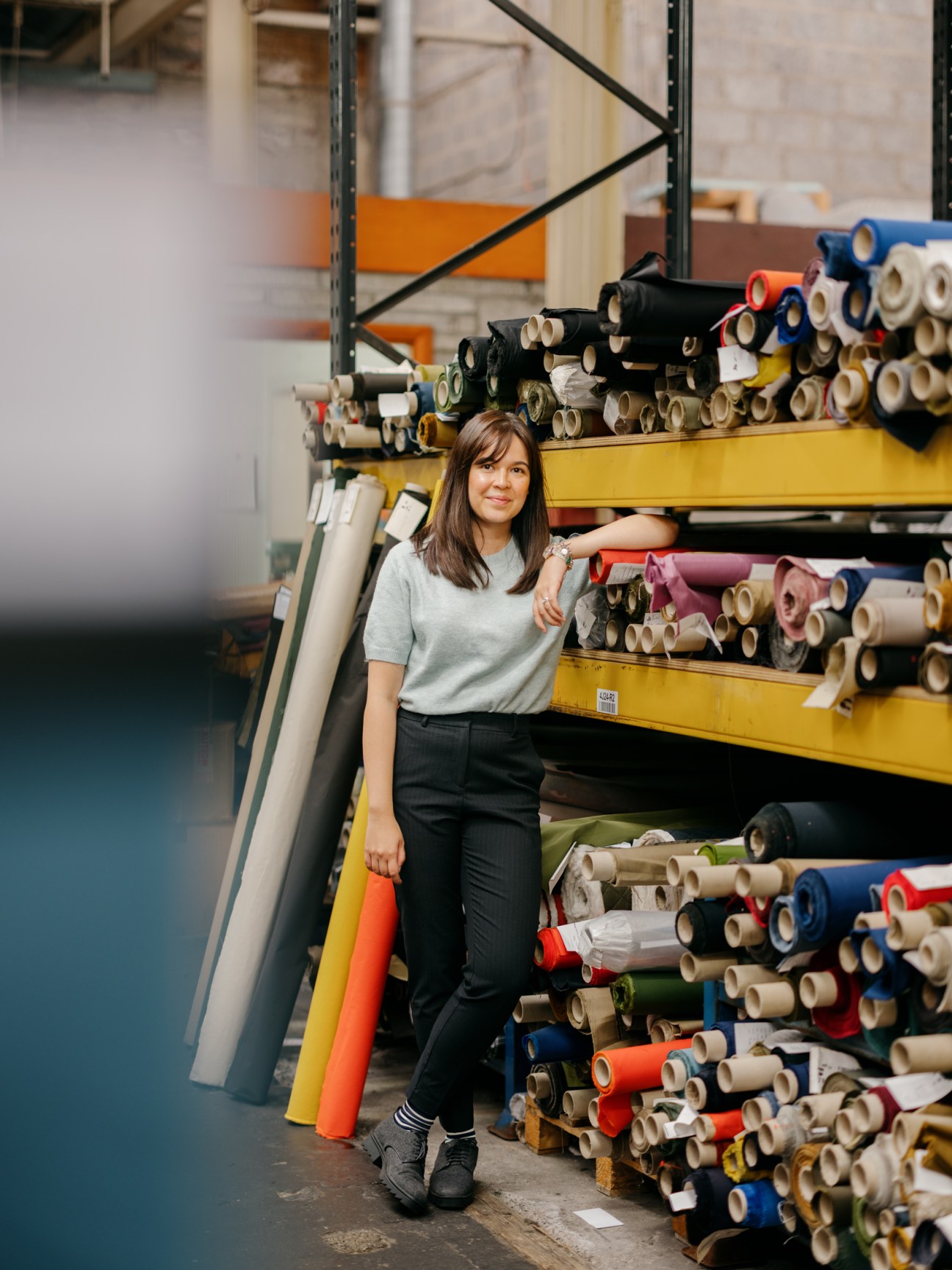 Woman with brown hair in light blue sweater and black pants standing next to shelves of colorful fabric rolls in warehouse