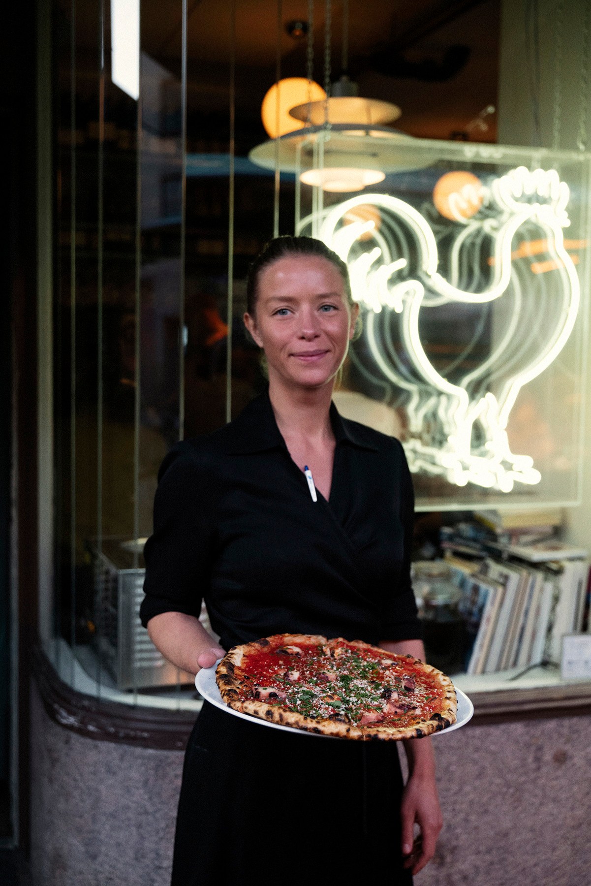 Woman in black holding a pizza outside restaurant with neon sign