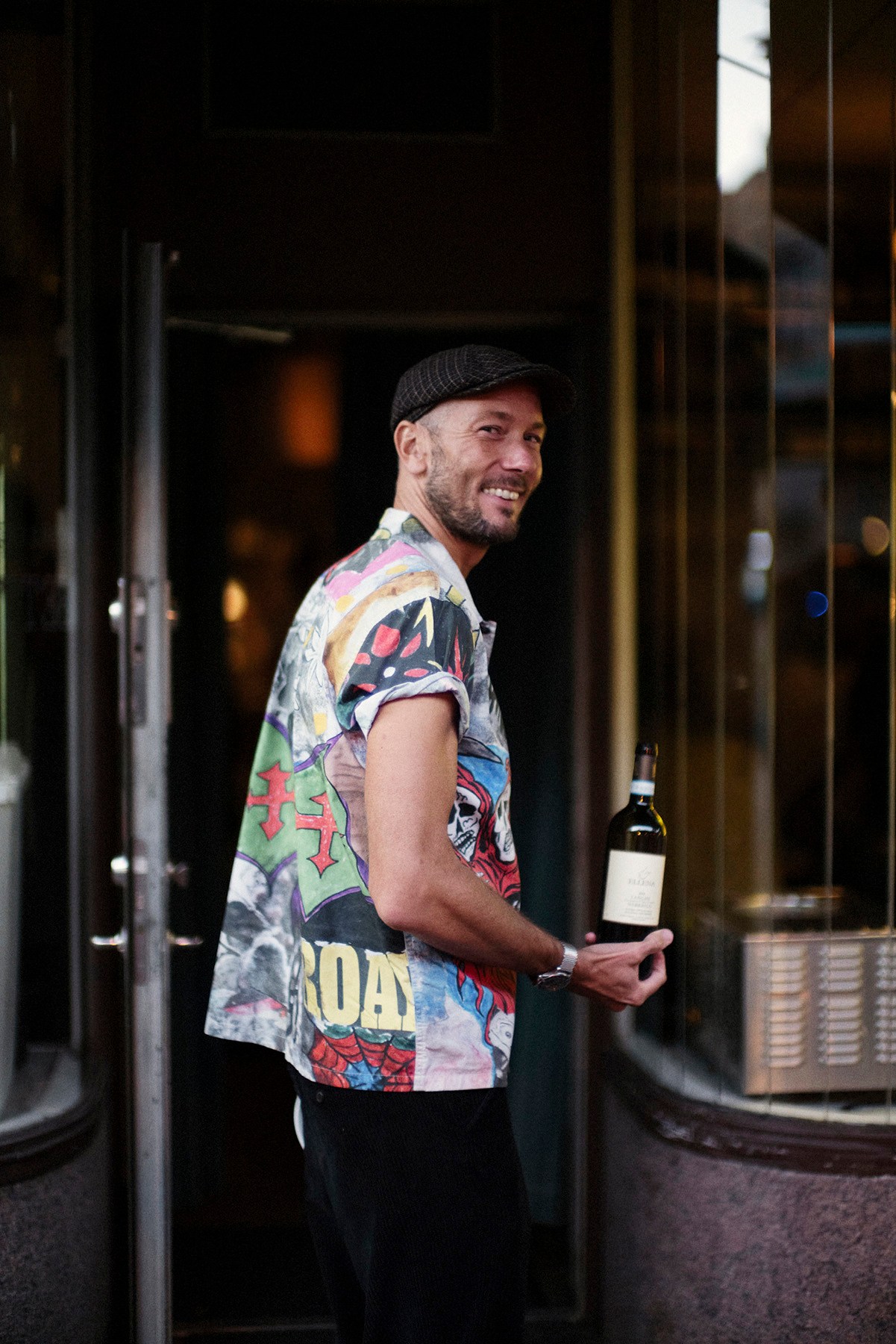 Man in colorful graphic t-shirt and flat cap holding wine bottle outside restaurant entrance