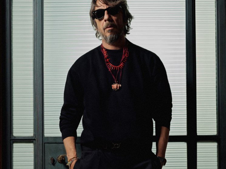Pierpaolo Piccioli in black sweater and sunglasses standing by window with venetian blinds