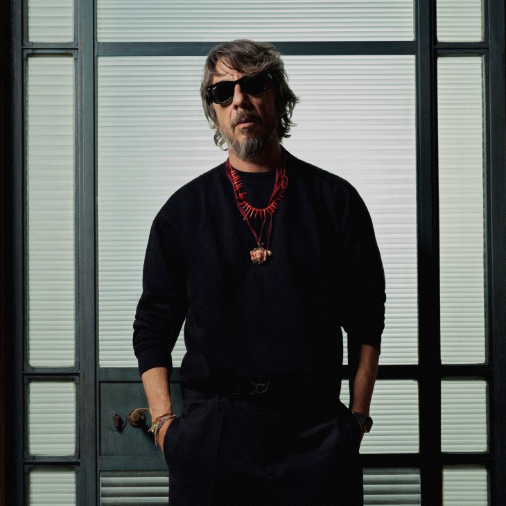 Pierpaolo Piccioli in black sweater and sunglasses standing by window with venetian blinds