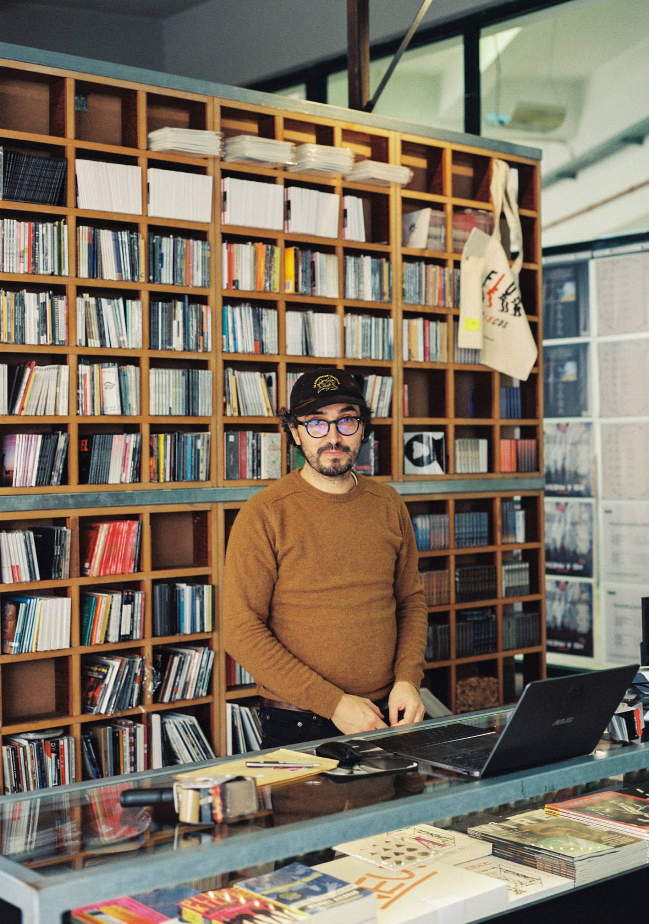 Man wearing glasses and mustard sweater stands at desk with laptop in front of wooden shelving unit filled with CDs and vinyl records.