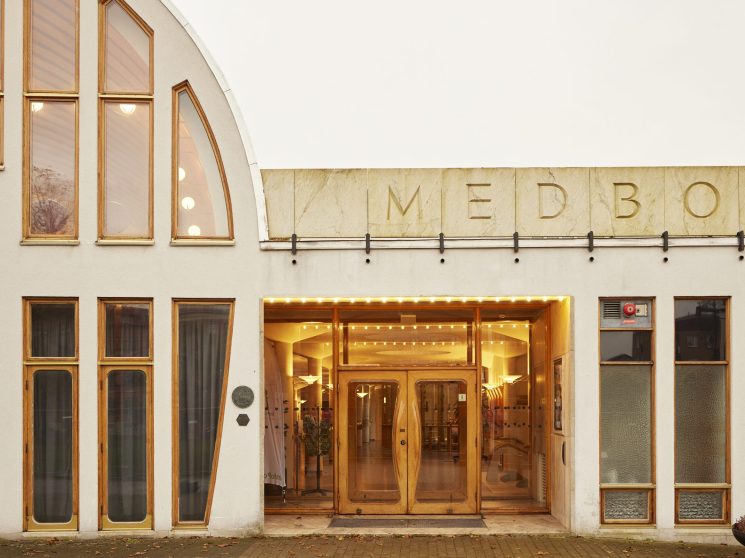 White modernist building exterior with wooden-framed windows and illuminated entrance displaying 'MEDBOR' lettering