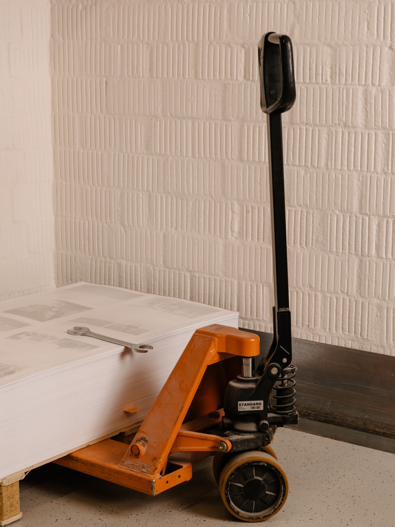 An orange and black hand pallet jack with printed pages stacked on its platform against a white brick wall.