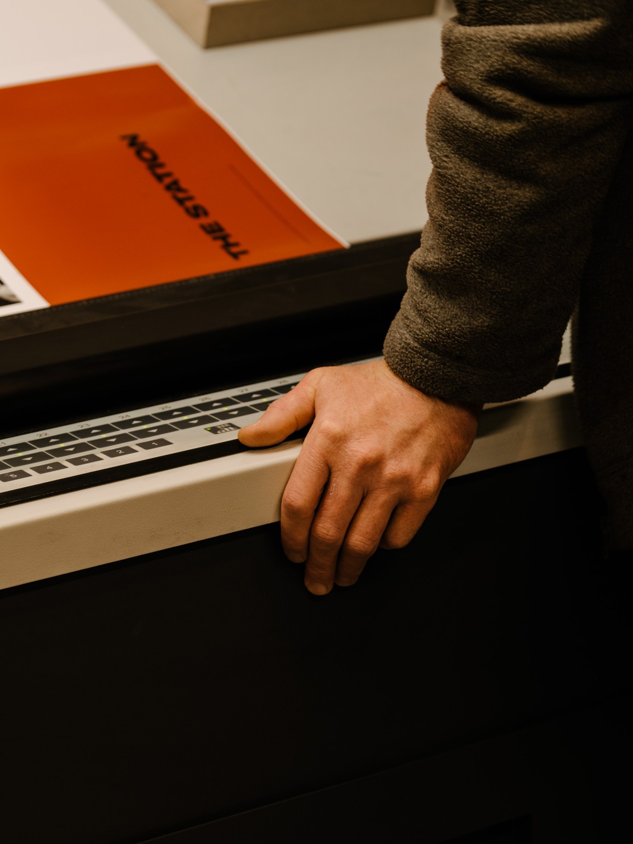 A hand rests on a printing press screen displaying black ink density patterns with an orange book visible in the background.