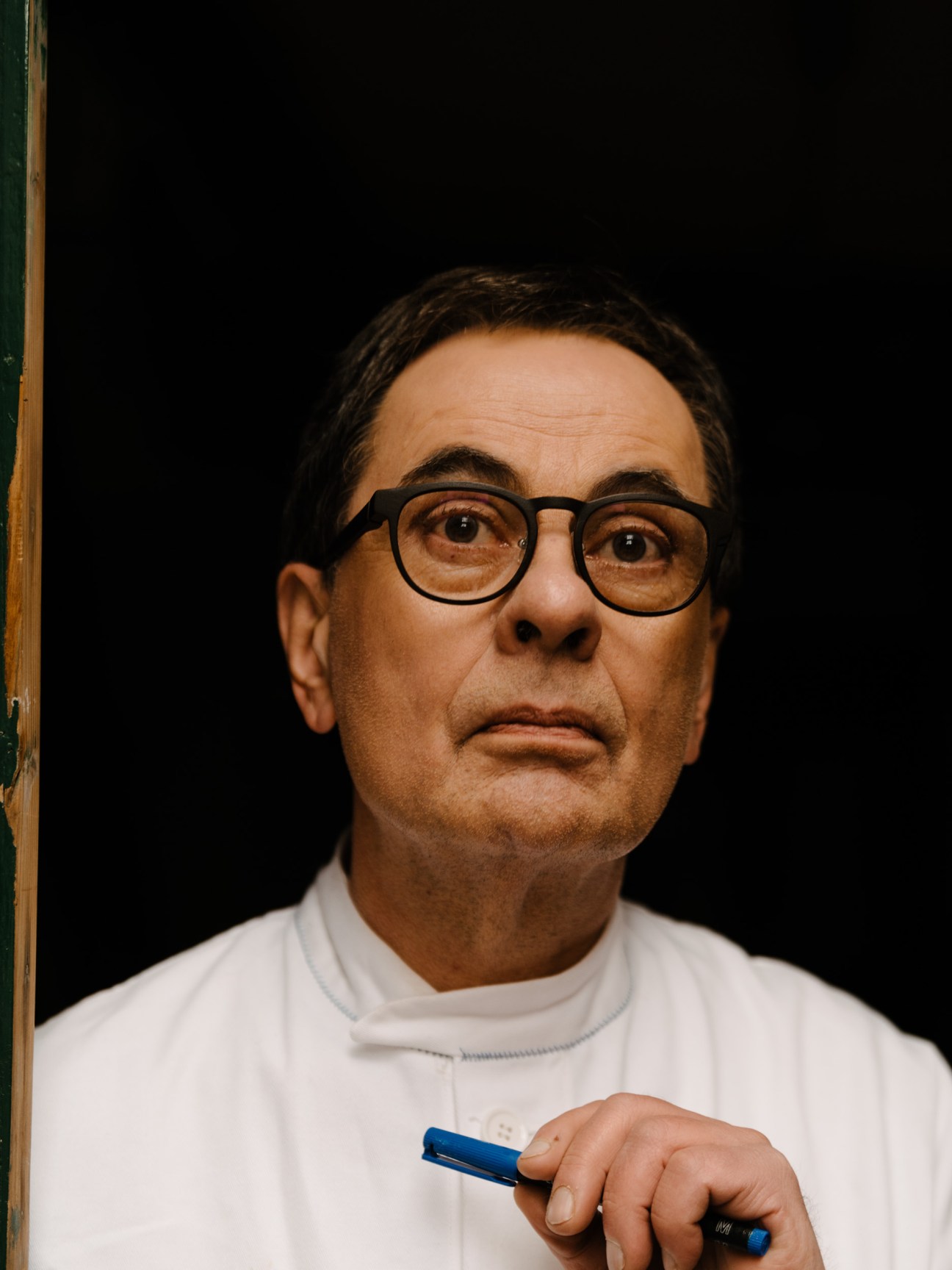 Publisher Gerhard Steidl wears glasses and a white apron while holding a blue pen against a black background.