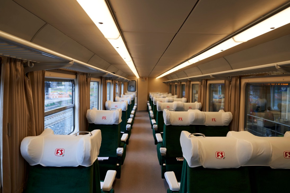 Interior of refurbished train car showing rows of green seats with white covers bearing FS logo and large windows