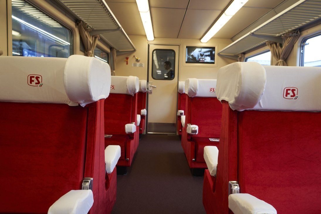 Interior view of L'Arlecchino train showing red and white first-class seats with white headrest covers and FS branding