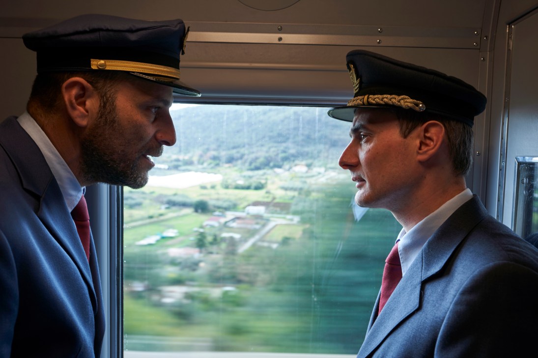 Two train conductors in vintage 1960s railway uniforms with peaked caps face each other aboard L'Arlecchino