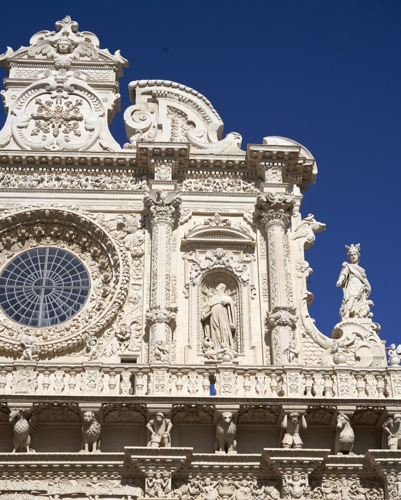 Ornate baroque stone facade with intricate carved decorations, rose window, statues and columns against blue sky