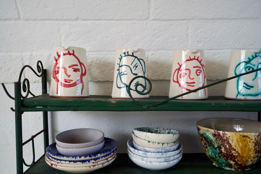 White ceramic mugs with colorful hand-drawn face illustrations and ceramic bowls stacked on green metal shelving