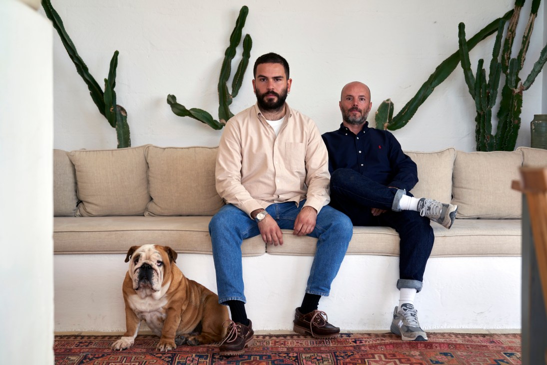 Two men and a bulldog sitting on a beige sofa with large cacti behind them in a modern white interior