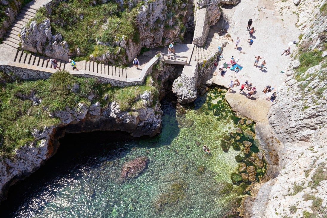 Aerial view of Porto Ciolo Beach showing dramatic limestone cliffs, turquoise water, and tourists on stone platforms