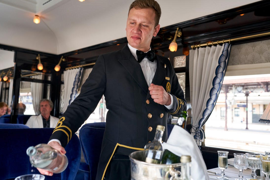 Uniformed steward in navy jacket with gold trim pouring champagne in elegant train dining car with blue seating
