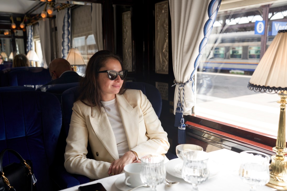 Woman in cream blazer and sunglasses sits at elegantly set dining table aboard luxury train looking out window