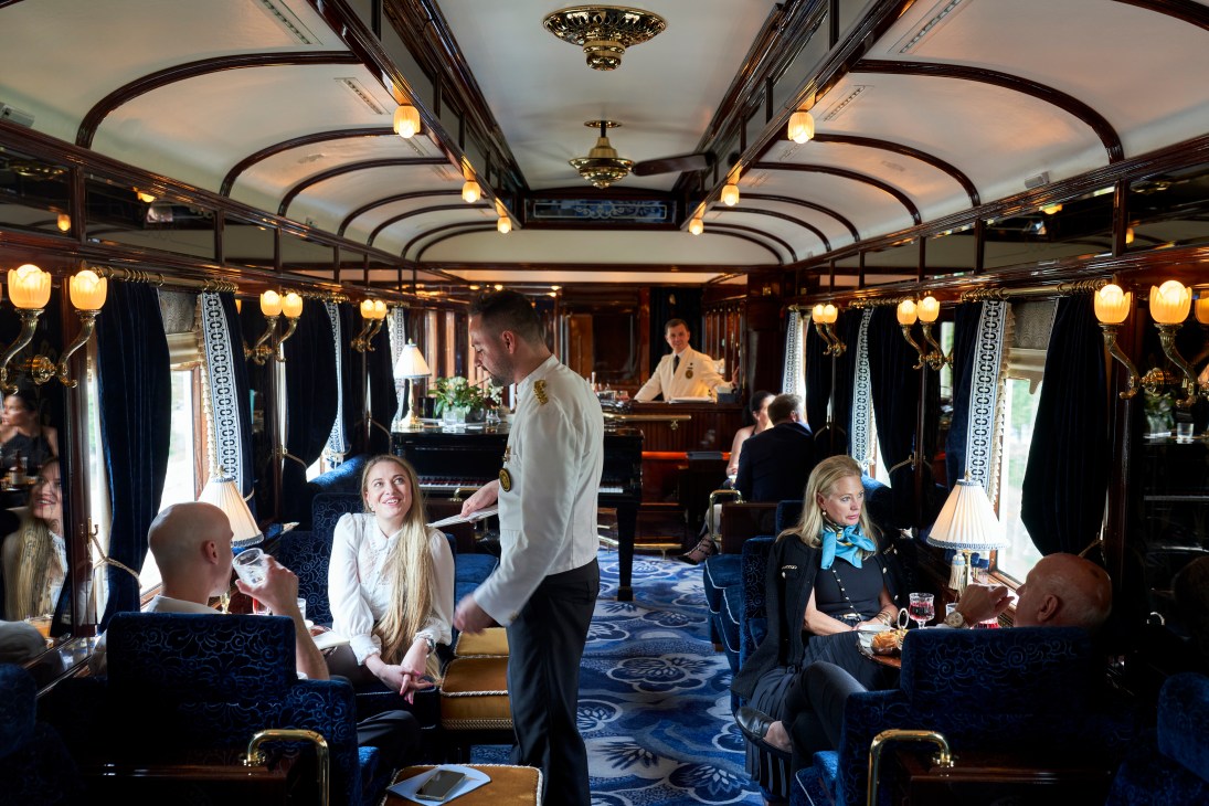 Passengers enjoying drinks in an ornate train bar car with blue upholstery, warm lighting and classic decor