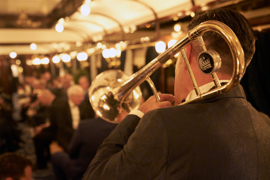 A musician in a dark suit plays a brass trombone in a warmly lit train car with string lights overhead