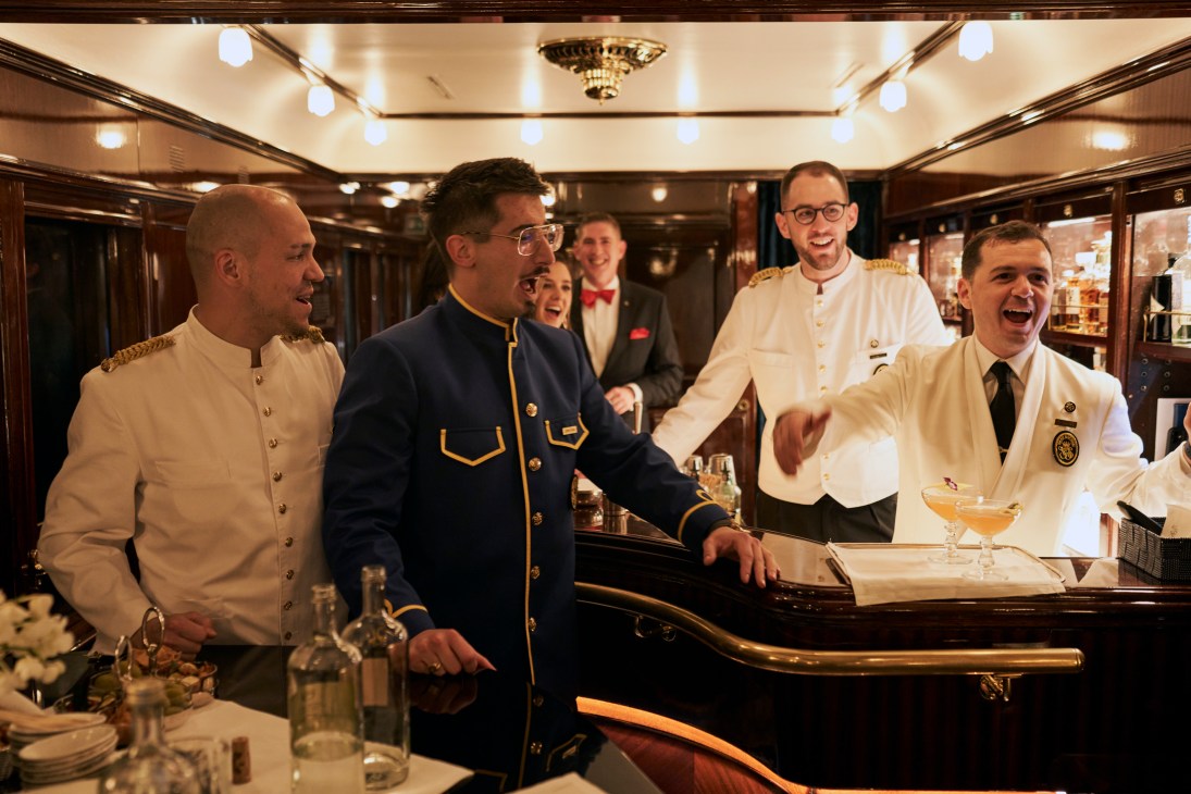 Train staff and passengers singing together in an elegant bar car with wood paneling and brass fixtures