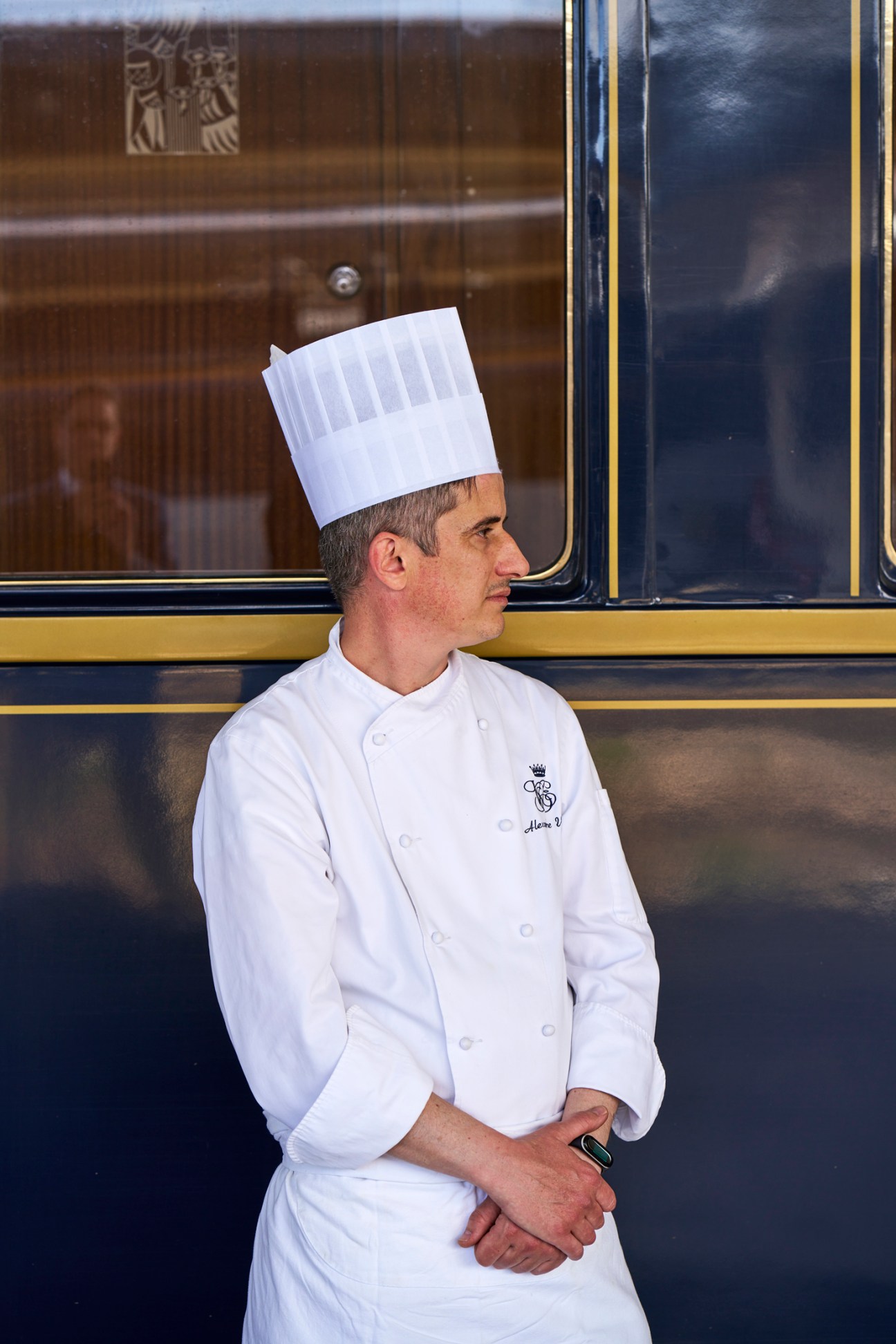 Chef in white uniform and traditional toque hat standing beside luxury train car with blue and gold exterior