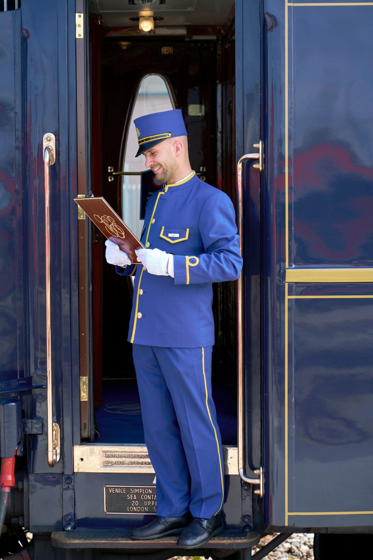 Train conductor in blue uniform with gold trim checking passenger list while standing in doorway of Orient Express carriage
