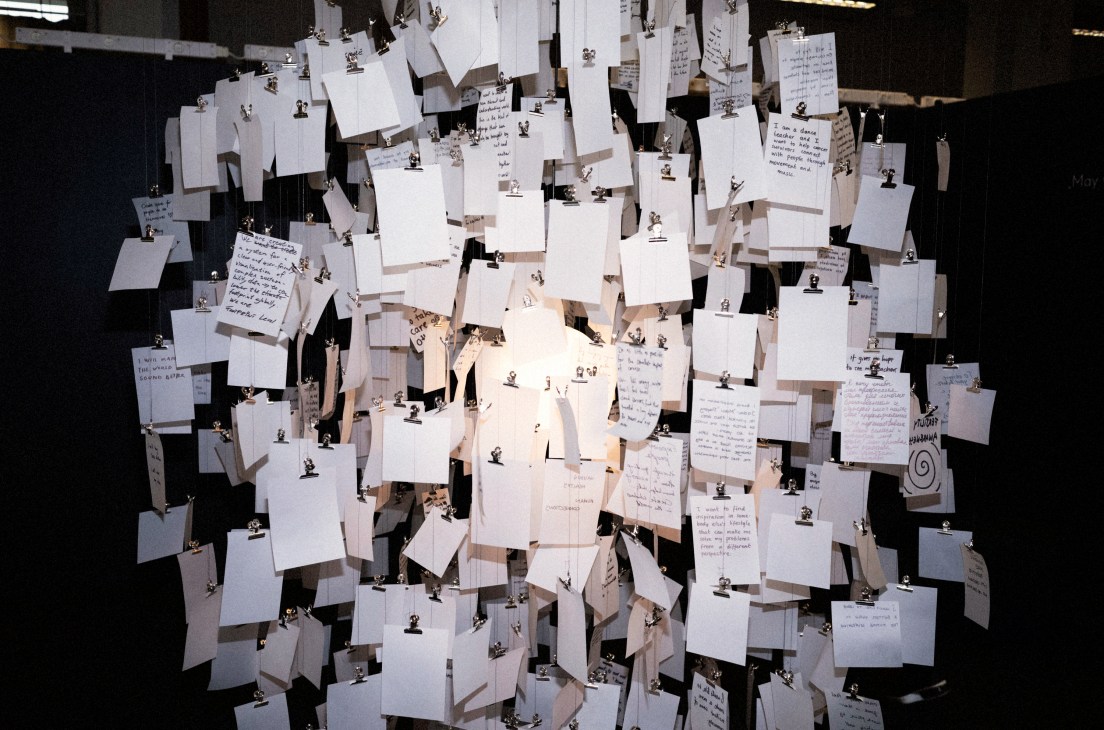 Many white papers with handwritten text hanging from metal clips attached to strings or wires