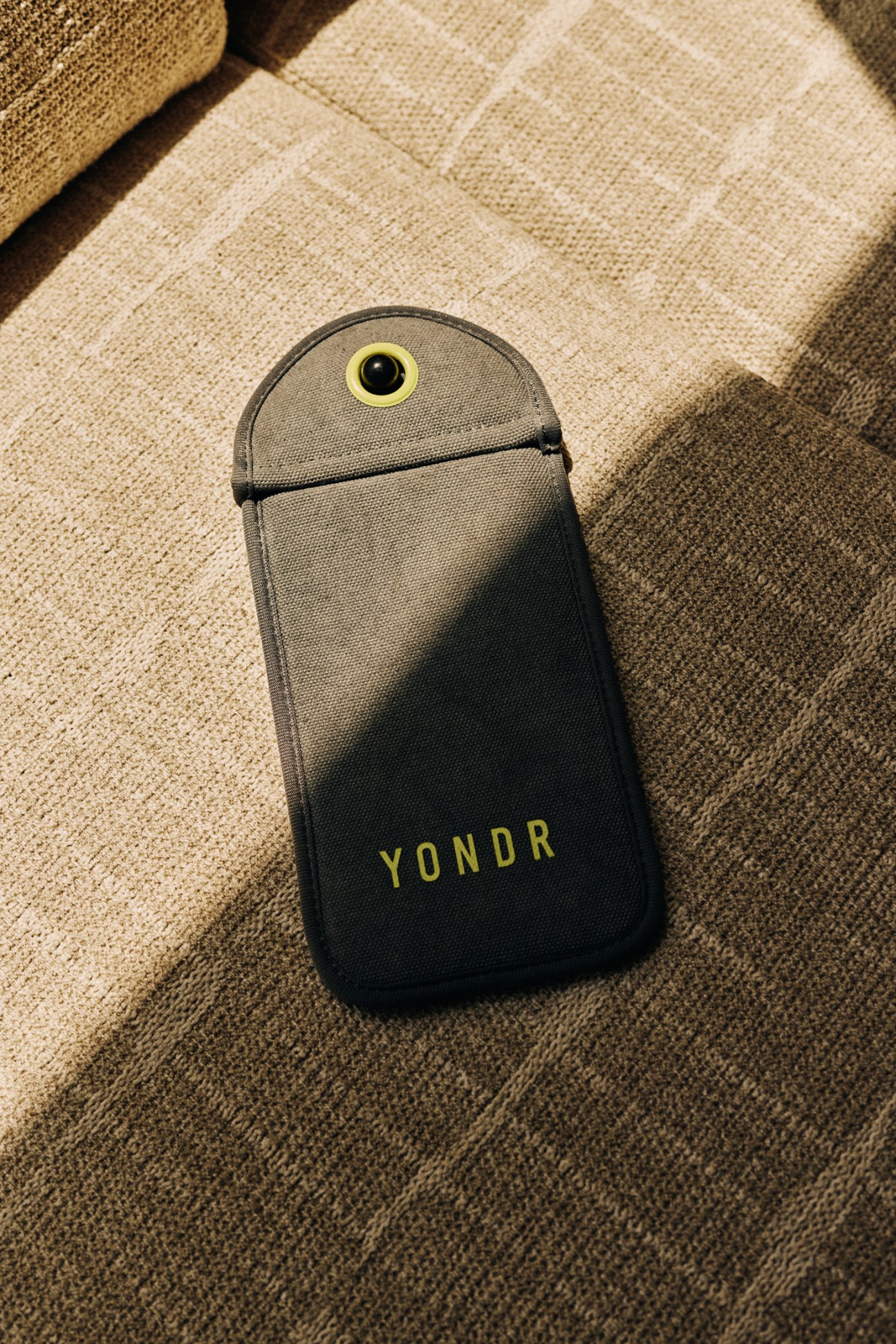 A gray and black Yondr phone pouch with yellow circular lock mechanism lying on a beige textured couch