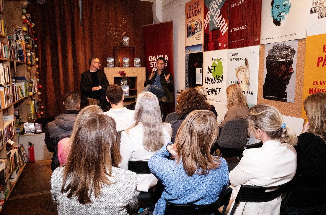 Audience seated on floor listening to two men speaking at bookstore event with book displays on walls