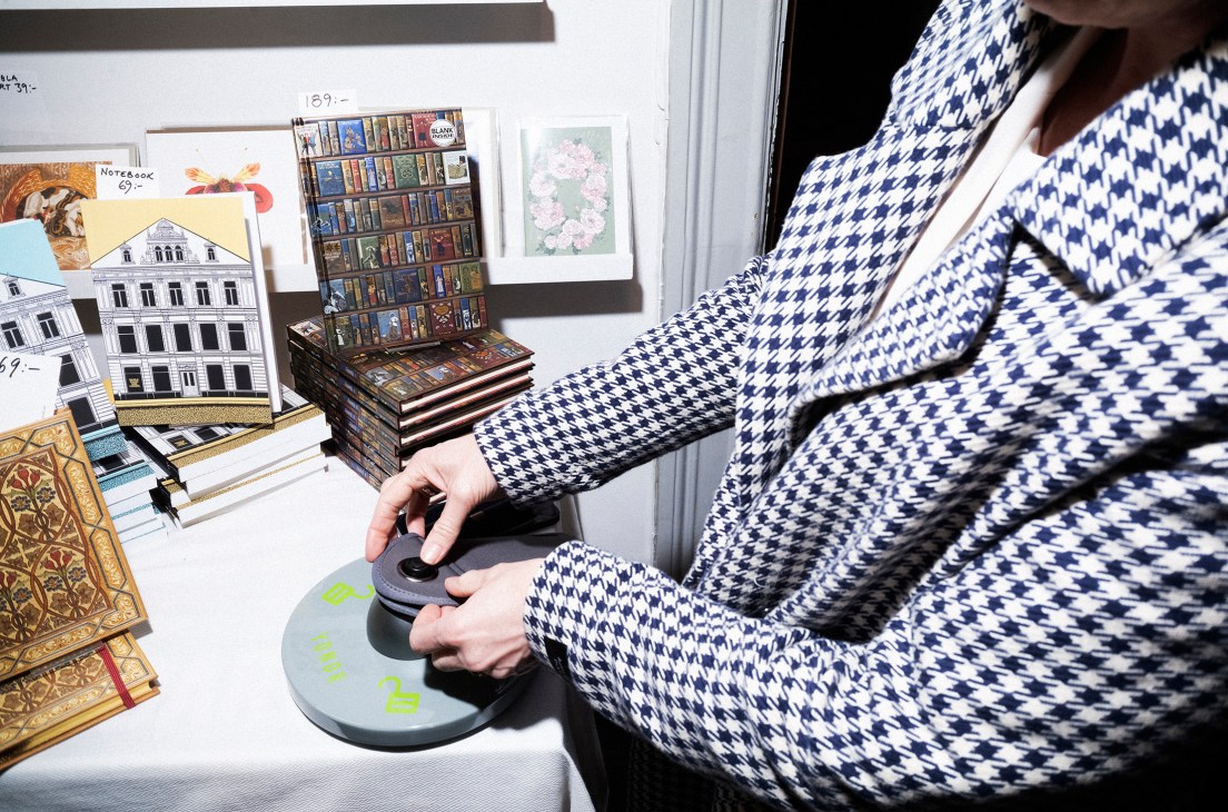 Person in blue and white houndstooth shirt unlocking a Yondr device to retrieve their phone from a pouch