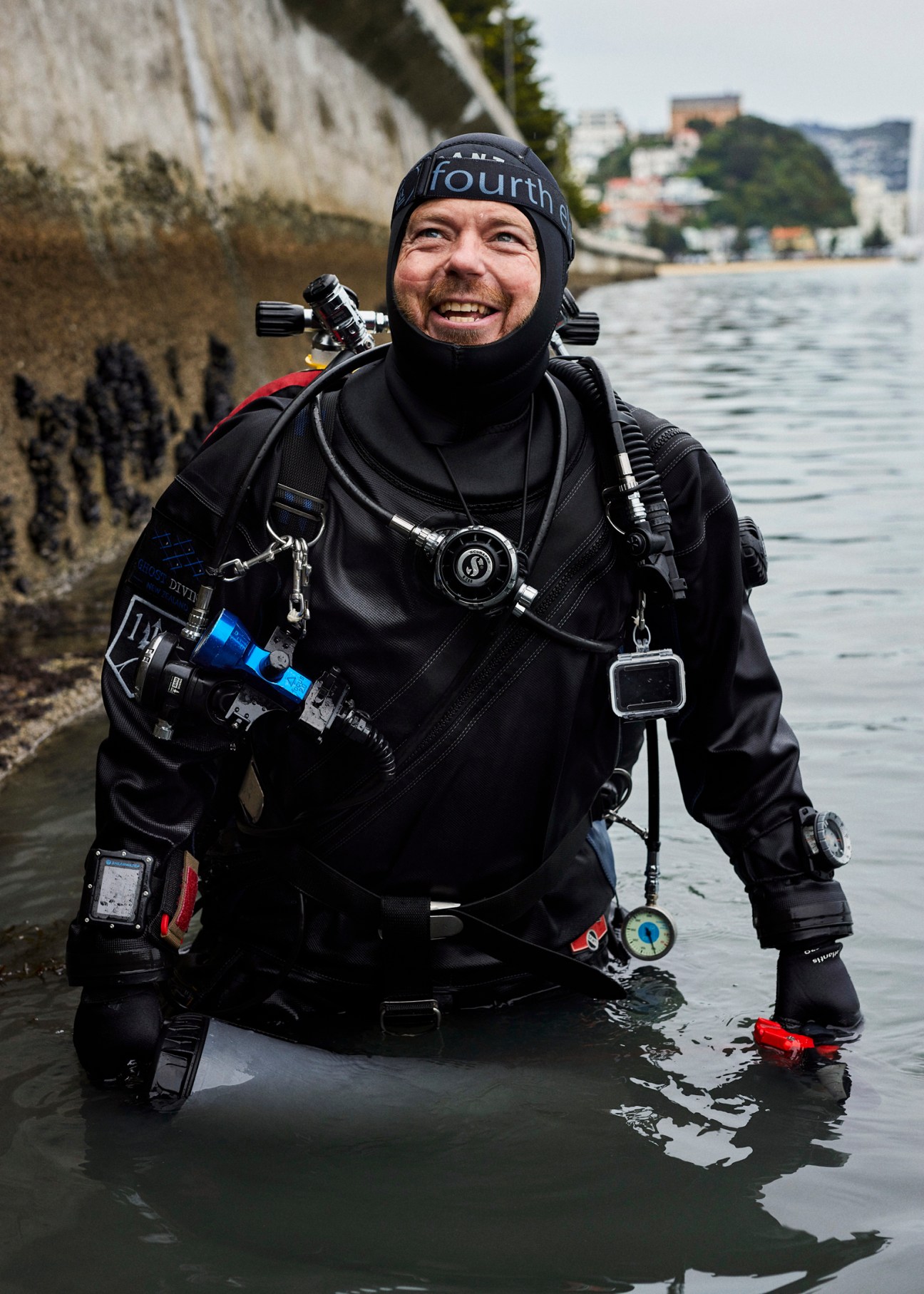 A smiling scuba diver in full gear stands in shallow water, equipped with underwater cameras and diving equipment