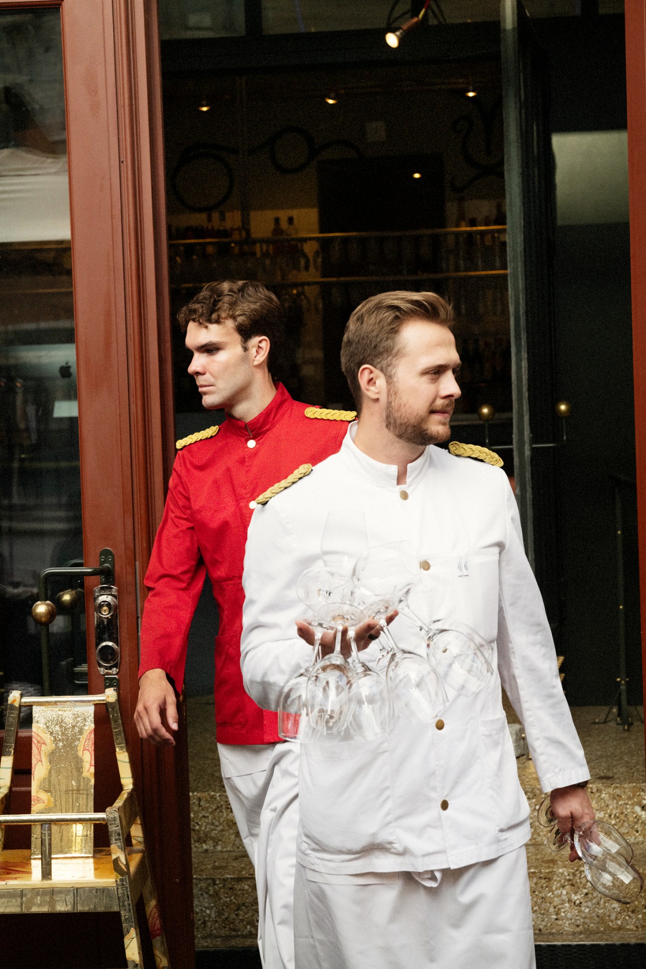 Two men in formal uniforms, one in red with gold trim and one in white chef's coat, standing outside restaurant