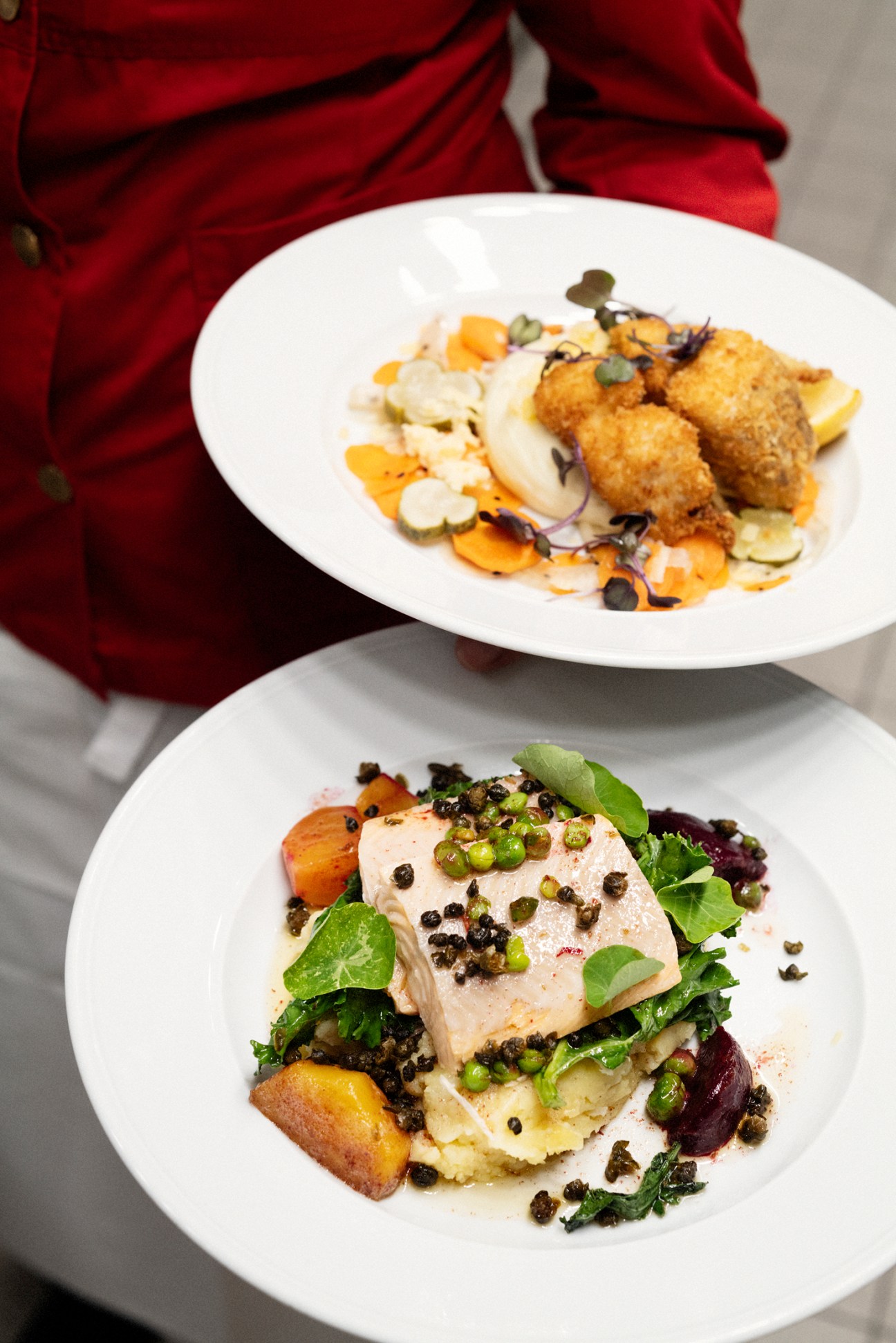Two white plates of gourmet food - one with breaded items and vegetables, another with fish and greens