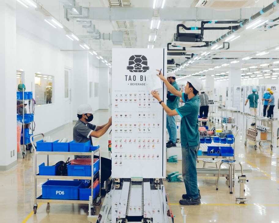 Workers in teal uniforms assembling vending machines on a factory production line