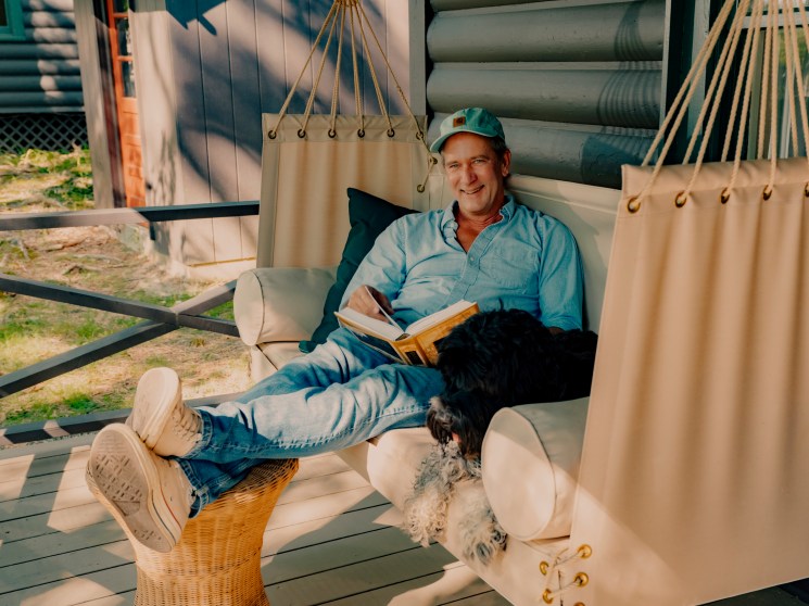 Man in light blue shirt and cap reading book in hammock with small black and white dog on wooden deck