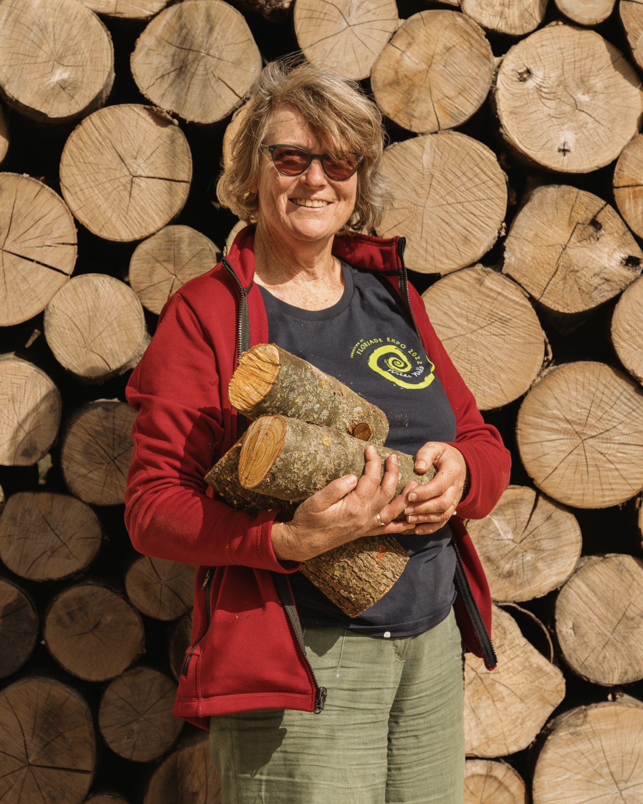 Woman in red jacket and sunglasses holding firewood logs against stacked wood background